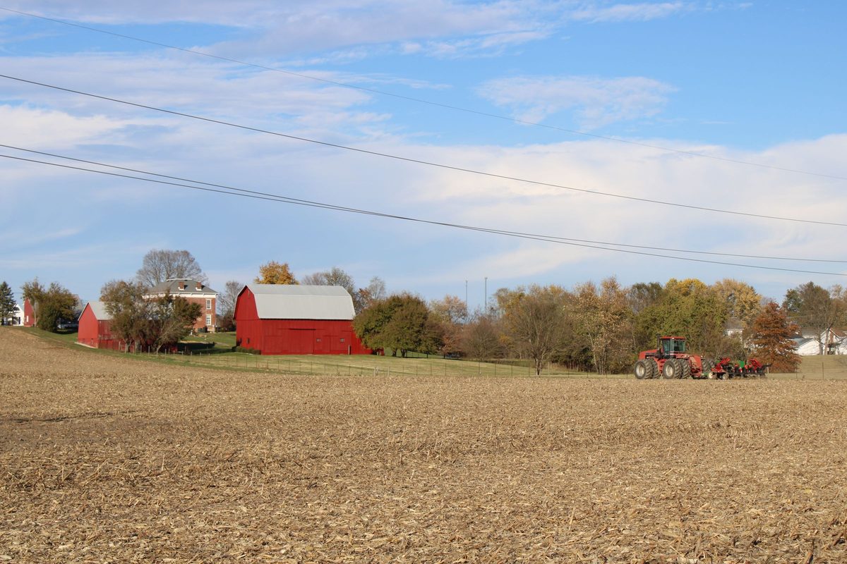 Red barns and a brick farmhouse beyond a harvested field with a tractor